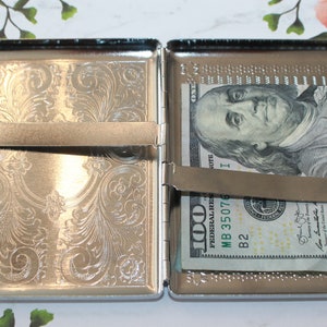 May include: Open silver-toned metal cigarette case with an ornate floral design. The case is open, revealing a one-hundred-dollar bill secured by a metal clip. The interior of the case has a decorative border.