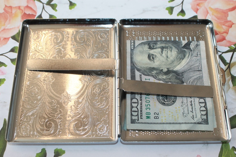 May include: Open silver-toned metal cigarette case with an ornate floral design. The case is open, revealing a one-hundred-dollar bill secured by a metal clip. The interior of the case has a decorative border.