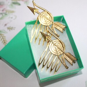 May include: Two gold-toned hair combs featuring a decorative design of a bow and arrow crossing over a circular target. The hair combs are presented inside a small, rectangular, bright green cardboard box with white foam padding inside.