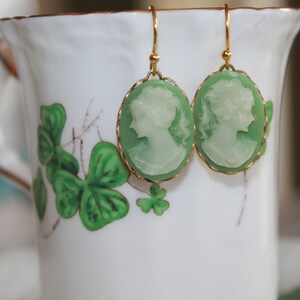 May include: A pair of green cameo earrings with gold accents. The earrings are shaped like ovals and feature a white cameo of a woman's profile. The earrings are hanging from a white cup with a green shamrock design.