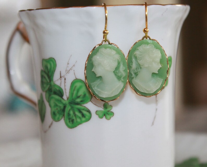 May include: A pair of green cameo earrings with gold accents. The earrings are shaped like ovals and feature a white cameo of a woman's profile. The earrings are hanging from a white cup with a green shamrock design.