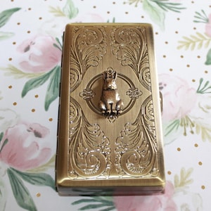 May include: A rectangular, antique brass-toned metal case featuring an intricate embossed scrollwork pattern on the lid. A small, three-dimensional rabbit charm is centred on the top surface, surrounded by a circular frame within the metal design.
