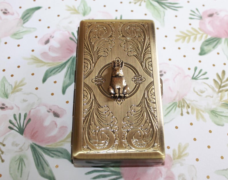 Puede incluir: Un estuche rectangular de metal en tono lat&oacute;n antiguo con un intrincado patr&oacute;n de volutas en relieve en la tapa. Un peque&ntilde;o dije de conejo tridimensional est&aacute; centrado en la superficie superior, rodeado por un marco circular en el dise&ntilde;o.