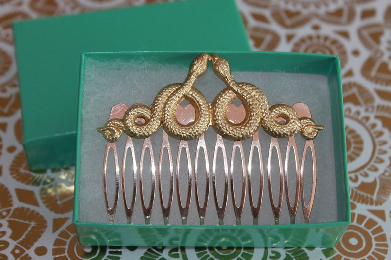 May include: A gold hair comb with a decorative design of two snakes intertwined. The comb has 10 teeth and is in a teal blue box.