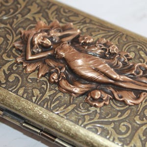 May include: A vintage-style metal cigarette case with an ornate design. The case features a raised, copper-colored image of a woman reclining in a floral setting.