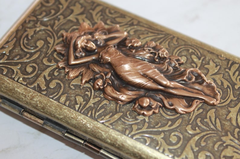 May include: A vintage-style metal cigarette case with an ornate design. The case features a raised, copper-colored image of a woman reclining in a floral setting.