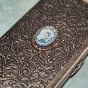 Puede incluir: Una caja met&aacute;lica rectangular con un dise&ntilde;o floral ornamentado. Un peque&ntilde;o camafeo ovalado con la cabeza de un carnero y la palabra "ARIES" est&aacute; centrado en la caja. El camafeo es azul y blanco, con el texto "MAR 21-APR 19".
