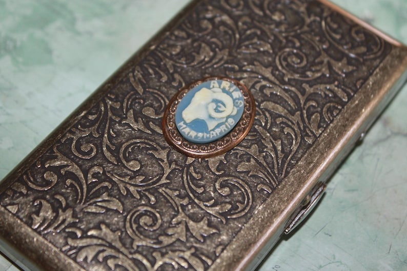 Puede incluir: Una caja met&aacute;lica rectangular con un dise&ntilde;o floral ornamentado. Un peque&ntilde;o camafeo ovalado con la cabeza de un carnero y la palabra "ARIES" est&aacute; centrado en la caja. El camafeo es azul y blanco, con el texto "MAR 21-APR 19".