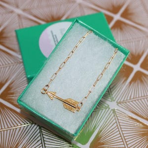 May include: A gold-toned necklace featuring a horizontal arrow pendant attached to a paperclip-style chain. The necklace is presented inside a bright green rectangular gift box lined with white padding, showcasing the metallic finish clearly.