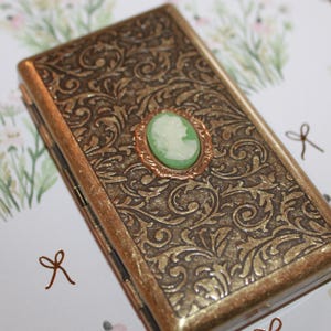 May include: A gold-colored metal cigarette case with an ornate floral design. The case has a hinged lid and a small green cameo with a white profile image in the center.