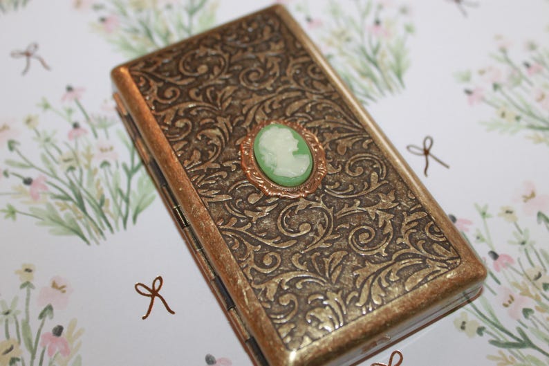May include: A gold-colored metal cigarette case with an ornate floral design. The case has a hinged lid and a small green cameo with a white profile image in the center.