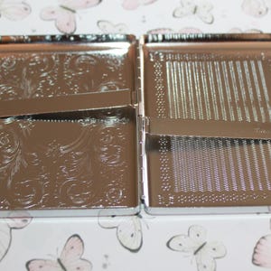 May include: Open, silver-colored cigarette case with an engraved floral design on one side and a lined pattern on the other. The case is rectangular and has a hinged opening. The background features a pattern of butterflies.