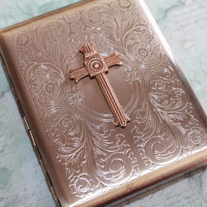 Puede incluir: Un estuche rectangular de metal en tono bronce con un intrincado patr&oacute;n floral en relieve en toda la superficie. Un prominente emblema de cruz tridimensional est&aacute; centrado en la parte frontal, mostrando las letras INRI en la parte superior.
