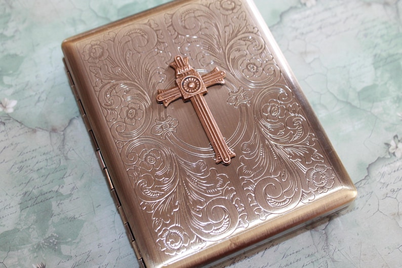 Puede incluir: Un estuche rectangular de metal en tono bronce con un intrincado patr&oacute;n floral en relieve en toda la superficie. Un prominente emblema de cruz tridimensional est&aacute; centrado en la parte frontal, mostrando las letras INRI en la parte superior.