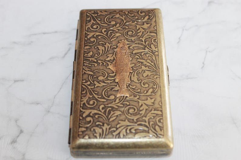 以下が含まれることがあります： 中央に魚のシルエットが描かれた、金色の金属製のタバコケース。ケースには、装飾的な花柄が施されています。