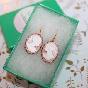May include: A pair of gold earrings with pink cameo pendants featuring a white silhouette of a woman's profile.
