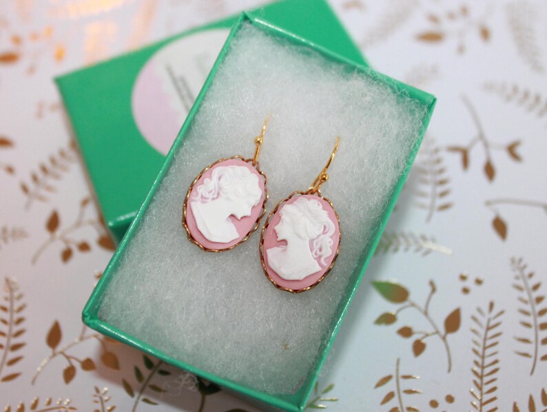 May include: A pair of gold earrings with pink cameo pendants featuring a white silhouette of a woman's profile.
