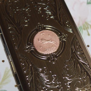 May include: A bronze-colored metal case with ornate floral scrollwork and a circular Aries emblem. The emblem features a ram and the word "ARIES". The case is rectangular and appears to be hinged.