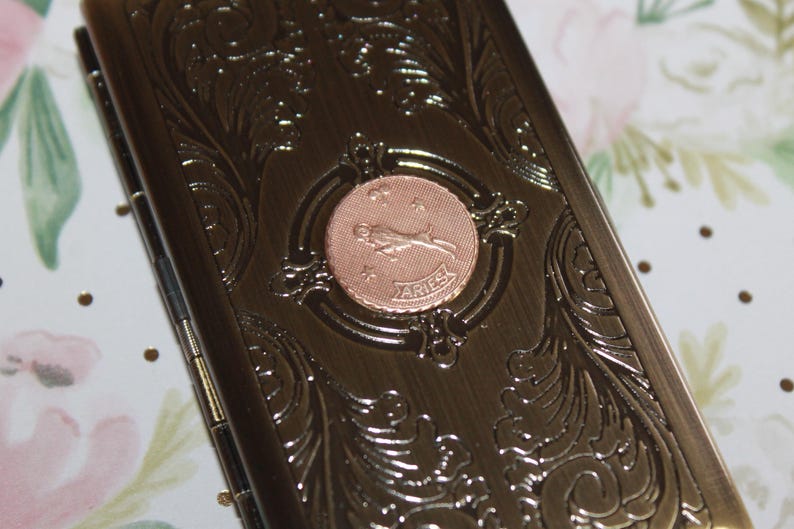 May include: A bronze-colored metal case with ornate floral scrollwork and a circular Aries emblem. The emblem features a ram and the word "ARIES". The case is rectangular and appears to be hinged.