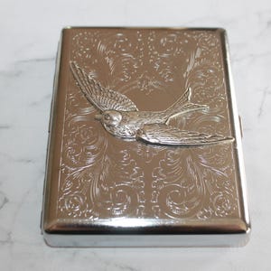 Puede incluir: Cajetilla de cigarrillos plateada con un patr&oacute;n floral en relieve y un dise&ntilde;o de p&aacute;jaro en 3D. La caja rectangular tiene bordes redondeados y un acabado brillante. El p&aacute;jaro est&aacute; en vuelo, agregando un elemento decorativo.