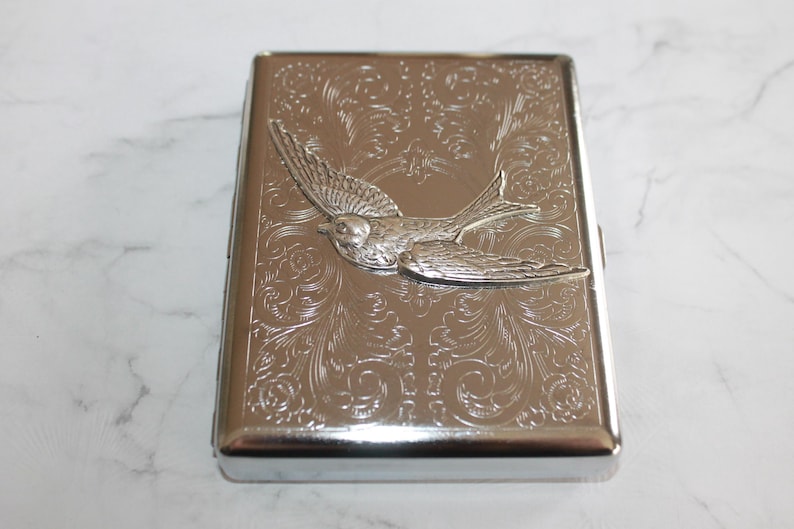 Puede incluir: Cajetilla de cigarrillos plateada con un patr&oacute;n floral en relieve y un dise&ntilde;o de p&aacute;jaro en 3D. La caja rectangular tiene bordes redondeados y un acabado brillante. El p&aacute;jaro est&aacute; en vuelo, agregando un elemento decorativo.
