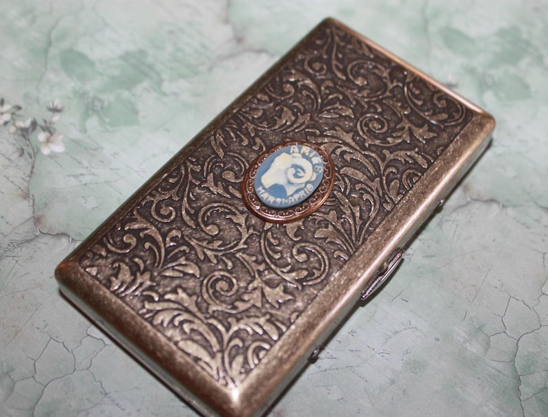 Puede incluir: Un estuche rectangular de metal de estilo antiguo con un intrincado dise&ntilde;o floral. Una peque&ntilde;a camafeo redondo azul y blanco est&aacute; fijado en la parte delantera. El estuche tiene un cierre de bisagra y un acabado met&aacute;lico.