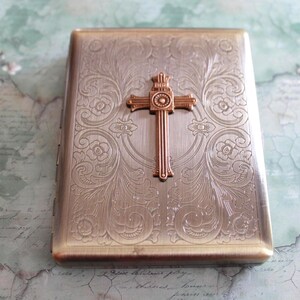 Puede incluir: Una pitillera rectangular de metal con acabado en bronce cepillado, que presenta un intrincado patr&oacute;n floral grabado en toda la superficie y un emblema de cruz en relieve de tono cobrizo centrado en la tapa frontal.