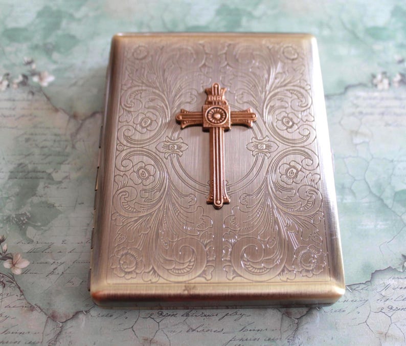 Puede incluir: Una pitillera rectangular de metal con acabado en bronce cepillado, que presenta un intrincado patr&oacute;n floral grabado en toda la superficie y un emblema de cruz en relieve de tono cobrizo centrado en la tapa frontal.