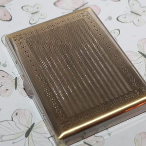 Puede incluir: Una pitillera rectangular de metal en tono dorado que presenta un panel central con finas l&iacute;neas onduladas verticales, bordeada por un marco decorativo con peque&ntilde;os detalles circulares perforados y acentos en forma de estrella en las esquinas.