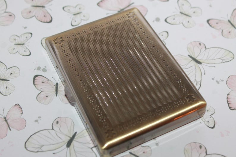 Puede incluir: Una pitillera rectangular de metal en tono dorado que presenta un panel central con finas l&iacute;neas onduladas verticales, bordeada por un marco decorativo con peque&ntilde;os detalles circulares perforados y acentos en forma de estrella en las esquinas.