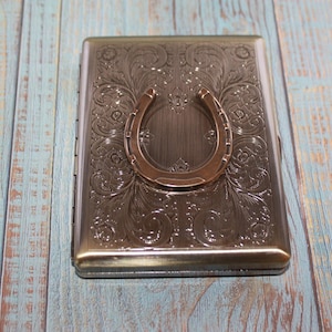 May include: A rectangular, metallic cigarette case with an embossed floral design and a bronze horseshoe accent. The case is a muted bronze color and sits on a weathered, blue-toned wooden surface.