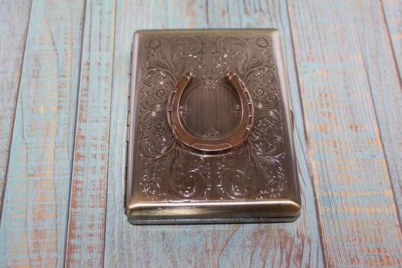 May include: A rectangular, metallic cigarette case with an embossed floral design and a bronze horseshoe accent. The case is a muted bronze color and sits on a weathered, blue-toned wooden surface.
