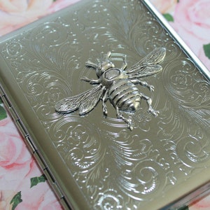 May include: Silver metal cigarette case with an embossed floral design and a 3D bee emblem. The rectangular case has a hinged lid. The bee is detailed with wings and legs. The case is approximately 10 cm long.