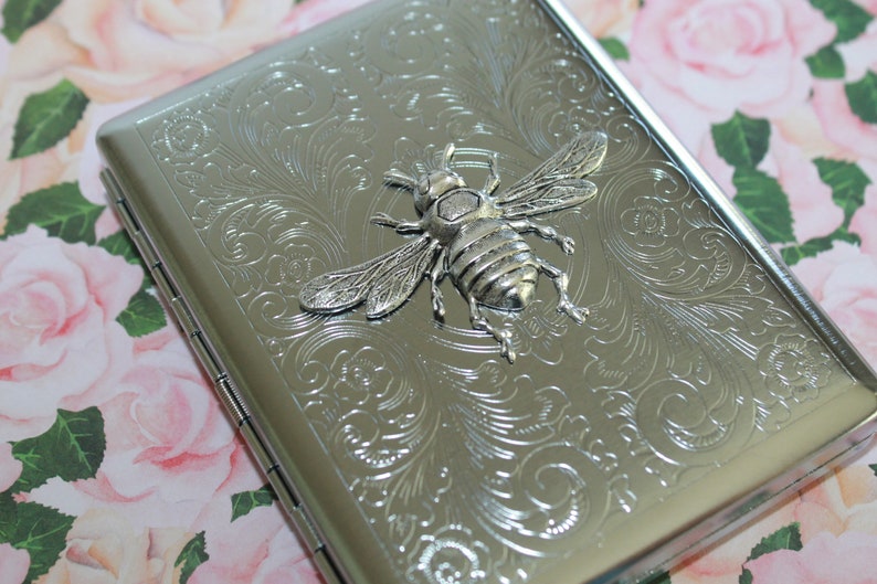 May include: Silver metal cigarette case with an embossed floral design and a 3D bee emblem. The rectangular case has a hinged lid. The bee is detailed with wings and legs. The case is approximately 10 cm long.