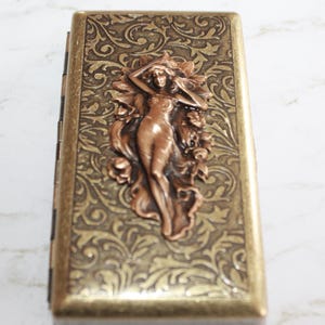 May include: A gold metal cigarette case with an ornate floral design and a copper-colored embossed figure of a woman.