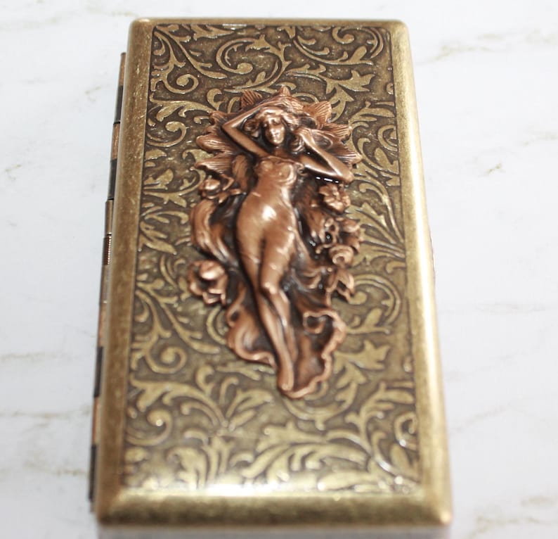 May include: A gold metal cigarette case with an ornate floral design and a copper-colored embossed figure of a woman.