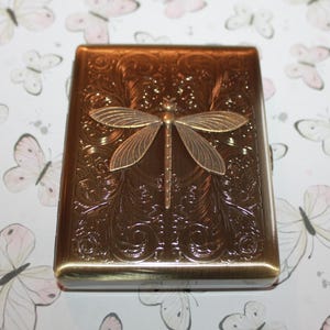 May include: A rectangular, bronze-coloured case with a detailed floral pattern and a dragonfly accent. The dragonfly is made of a lighter bronze metal and is centred on the case. The case has rounded edges.