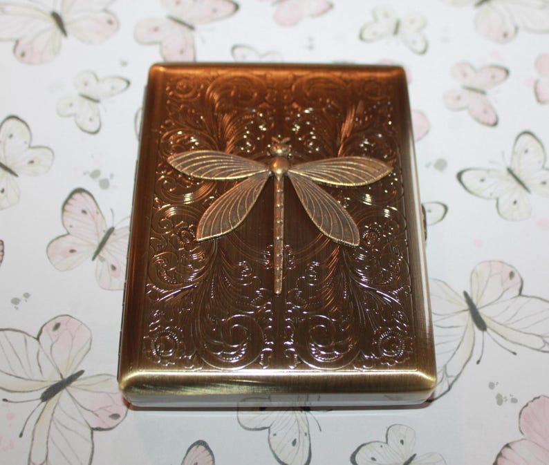 May include: A rectangular, bronze-coloured case with a detailed floral pattern and a dragonfly accent. The dragonfly is made of a lighter bronze metal and is centred on the case. The case has rounded edges.
