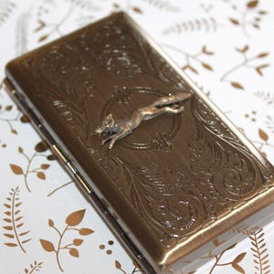 Puede incluir: Un estuche rectangular de metal en tono bronce con una figura de zorro en relieve centrada en la tapa. La superficie está decorada con intrincados patrones de volutas y hojas. El estuche tiene un lado con bisagras y un acabado metálico.