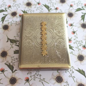 May include: A rectangular gold-toned metal case featuring an intricate engraved scrollwork pattern across the surface. A vertical row of seven small, raised, five-petaled flower embellishments is centered on the front of the metal container.