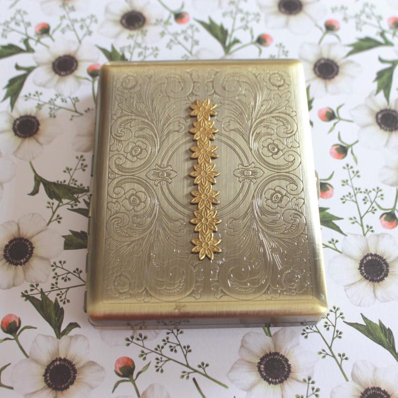 May include: A rectangular gold-toned metal case featuring an intricate engraved scrollwork pattern across the surface. A vertical row of seven small, raised, five-petaled flower embellishments is centered on the front of the metal container.
