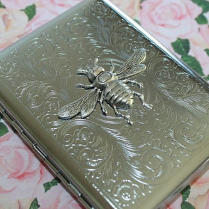 May include: Silver metal cigarette case with an embossed floral pattern and a 3D bee emblem. The case has a hinged lid and a rectangular shape. The bee is detailed with wings and body segments.