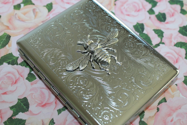 May include: Silver metal cigarette case with an embossed floral pattern and a 3D bee emblem. The case has a hinged lid and a rectangular shape. The bee is detailed with wings and body segments.