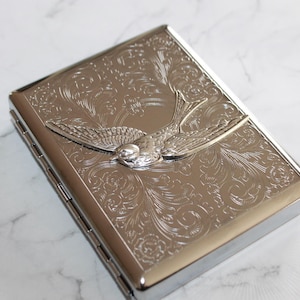 Könnte beinhalten: Eine silberfarbene Zigarettenetui aus Metall mit einem gravierten Vogelmotiv. Die rechteckige Hülle hat einen Klappdeckel und abgerundete Kanten. Der Vogel ist im Flug dargestellt, mit ausgebreiteten Flügeln. Die Hülle ist hochglanzpoliert und reflektiert das Licht.