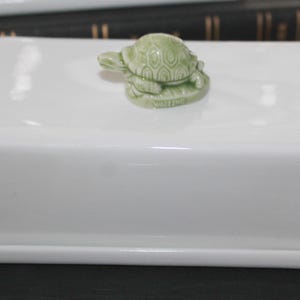 May include: White ceramic butter dish with a green ceramic turtle figurine on top. The dish has a raised rim and a base.