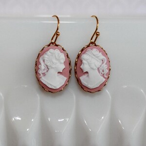 May include: A pair of gold earrings with pink cameo portraits of women. The cameos are oval shaped and have a white raised design.