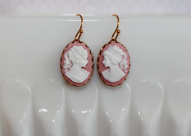 May include: A pair of gold earrings with pink cameo portraits of women. The cameos are oval shaped and have a white raised design.
