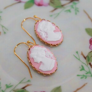 May include: A pair of pink cameo earrings with gold accents. The cameos feature a white profile of a woman's face.