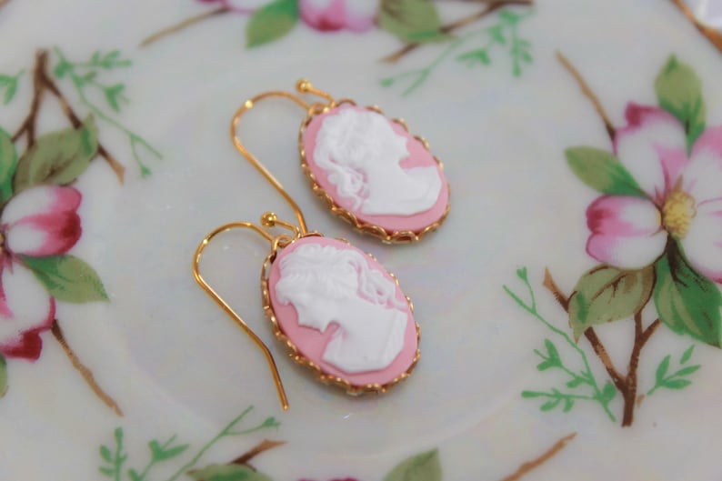 May include: A pair of pink cameo earrings with gold accents. The cameos feature a white profile of a woman's face.