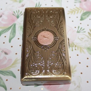 May include: A rectangular, antique brass-colored case with ornate floral and scrollwork designs. A circular copper-toned emblem in the center features a leaping figure and the word "ARIES". The case has a hinged lid and a clasp.